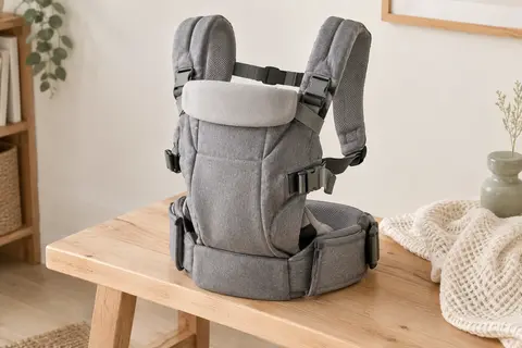 Porte-bébé convertible gris avec bretelles rembourrées, sangles réglables et appui-tête, posé sur un banc en bois clair.
