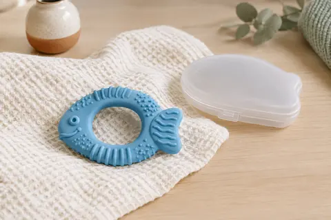 Anneau de dentition bleu Nuby en silicone texturé, en forme de poisson, posé près de son étui.